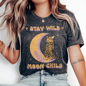 Könnte beinhalten: Ein dunkelgraues Heather-T-Shirt mit dem Text "Stay Wild Moon Child" in gelben und violetten Buchstaben. Hinter dem Text befindet sich ein gelber Halbmond mit einem violetten Blatt und einem darauf sitzenden Leoparden.