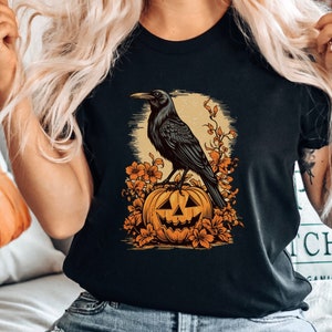 May include: Black t-shirt with a graphic of a crow perched on a jack-o'-lantern. The crow is black and the jack-o'-lantern is orange with a carved face. The background is a dark orange color with leaves and flowers.