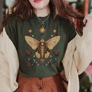 May include: A woman wearing a green t-shirt with a gold butterfly graphic and a brown corduroy skirt. The t-shirt features a celestial design with stars and moons.