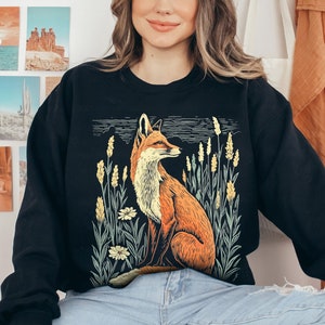 May include: Black sweatshirt with a graphic print of a fox in a field of wildflowers. The fox is orange and white and the flowers are yellow and green. The text "Forest Fables" is printed in the top right corner of the image.