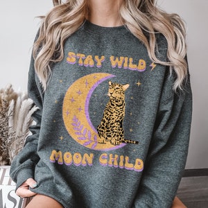 Puede incluir: Sudadera gris oscuro jaspeada con un gráfico de un leopardo sentado en una luna creciente. El texto "Stay Wild Moon Child" está impreso en morado y amarillo en la sudadera.