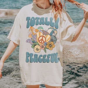 May include: A white t-shirt with a retro graphic design featuring the text "Totally Peaceful" in blue letters with a peace sign and flowers. The shirt is a relaxed fit and has a crew neck.
