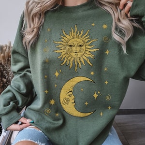 Puede incluir: Una sudadera verde con un sol amarillo y una luna creciente blanca. El sol y la luna tienen caras y están rodeados de pequeñas estrellas amarillas.