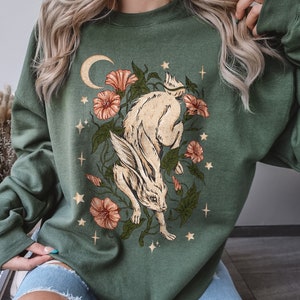 May include: A green sweatshirt with a white rabbit illustration surrounded by flowers and stars. The rabbit is jumping and has a crescent moon behind it.