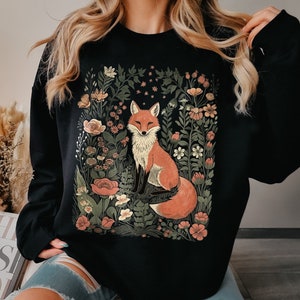 May include: Black sweatshirt with a graphic print of a fox in a floral garden. The fox is orange and white with a bushy tail. The flowers are in shades of pink, purple, and green.