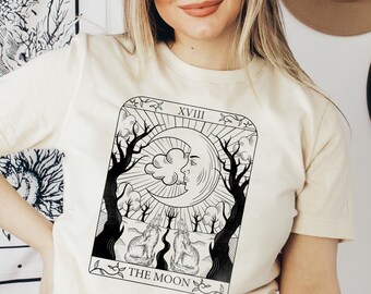 May include: A cream colored t-shirt with a black and white illustration of a tarot card. The card is the "The Moon" card and features a crescent moon with a face, two dogs, and trees. The card number, XVIII, is at the top of the card.