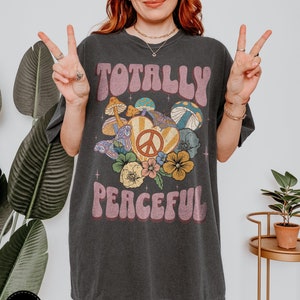 May include: An oversized dark grey t-shirt with a retro graphic design featuring the text "Totally Peaceful" in a groovy font. The graphic also includes a peace sign, mushrooms, butterflies, and flowers.