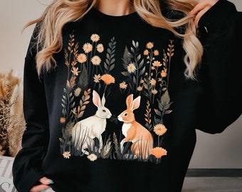May include: Black sweatshirt with a graphic print of two rabbits in a field of flowers. The rabbits are facing each other and appear to be interacting. The flowers are in shades of orange, yellow, and white.