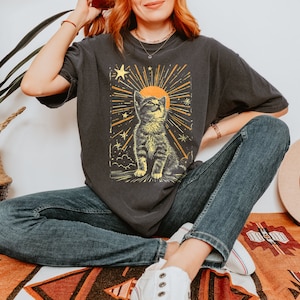 May include: A black t-shirt with a graphic of a cat looking up at a sun with stars and rays of light. The cat is brown and white and the sun is yellow. The text "Forest Fables" is in the top right corner of the image.