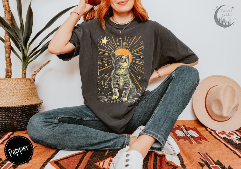 May include: A black t-shirt with a graphic of a cat looking up at a sun with stars and rays of light. The cat is brown and white and the sun is yellow. The text "Forest Fables" is in the top right corner of the image.