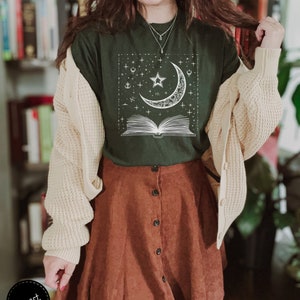 May include: A dark green t-shirt with a white graphic of a crescent moon, a star, and an open book with stars and other celestial symbols. The shirt is being worn by a person with a brown corduroy skirt and a cream cardigan.