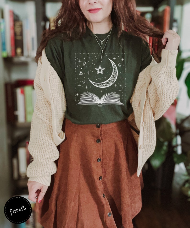 May include: A dark green t-shirt with a white graphic of a crescent moon, a star, and an open book with stars and other celestial symbols. The shirt is being worn by a person with a brown corduroy skirt and a cream cardigan.