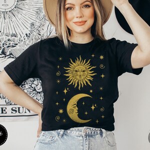 May include: A black t-shirt with a gold sun and moon design. The sun and moon have faces and are surrounded by stars. The text "Forest Fables" is visible in the lower right corner of the image.