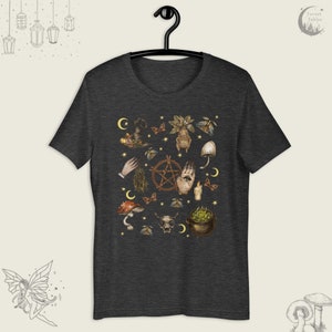 Puede incluir: Una camiseta gris oscuro jaspeada con un estampado de varios s&iacute;mbolos e im&aacute;genes de brujer&iacute;a, incluyendo un pentagrama, una mano con un ojo, setas, un caldero y una calavera. El estampado est&aacute; en tonos marrones, verdes y dorados.