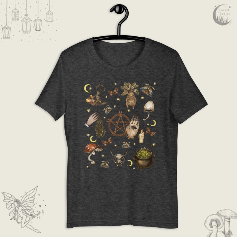 Puede incluir: Una camiseta gris oscuro jaspeada con un estampado de varios s&iacute;mbolos e im&aacute;genes de brujer&iacute;a, incluyendo un pentagrama, una mano con un ojo, setas, un caldero y una calavera. El estampado est&aacute; en tonos marrones, verdes y dorados.