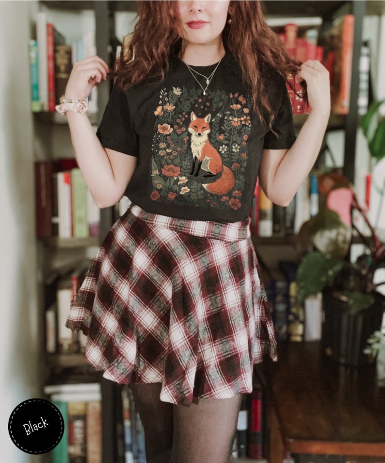 May include: Black t-shirt with a fox illustration surrounded by flowers. The person is wearing a plaid skirt in shades of red, white, and black. The skirt has a flared design. The image is taken indoors.