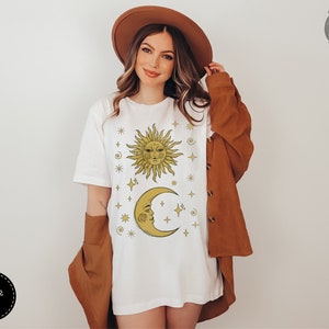 May include: White t-shirt with a gold sun and moon design with stars and other celestial symbols. The text "Forest Fables" is in the top right corner of the image.