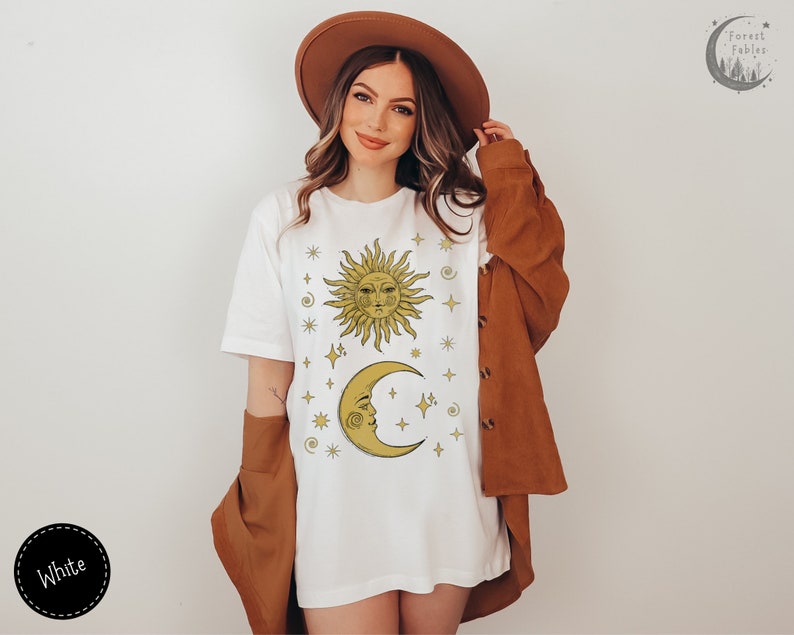 May include: White t-shirt with a gold sun and moon design with stars and other celestial symbols. The text "Forest Fables" is in the top right corner of the image.