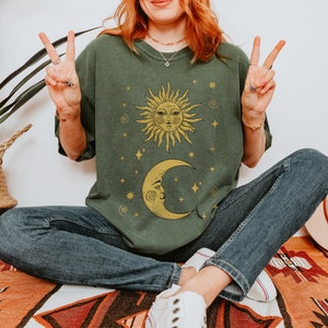 Puede incluir: Una camiseta verde con un dise&ntilde;o de sol amarillo y luna creciente. El sol y la luna tienen caras y est&aacute;n rodeados de estrellas. El texto "Forest Fables" est&aacute; en la esquina superior derecha de la imagen.
