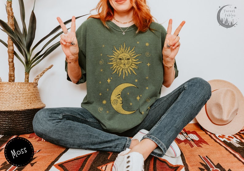Puede incluir: Una camiseta verde con un dise&ntilde;o de sol amarillo y luna creciente. El sol y la luna tienen caras y est&aacute;n rodeados de estrellas. El texto "Forest Fables" est&aacute; en la esquina superior derecha de la imagen.
