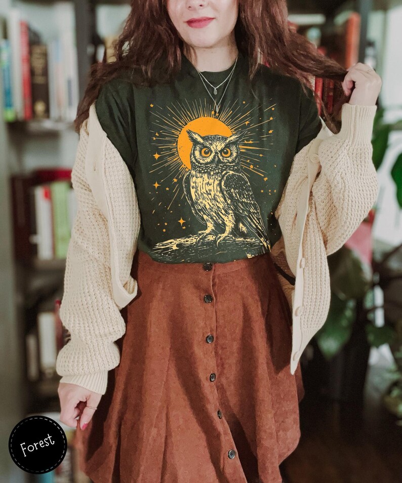 May include: A green t-shirt featuring a vintage illustration of an owl in front of a yellow sun and stars. The owl is perched on a branch. The shirt is worn with a cream cardigan and a brown corduroy skirt.