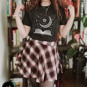 May include: Black t-shirt with a white graphic of a crescent moon, a star, and an open book with stars and other celestial symbols. The shirt is worn with a red and white plaid skirt.