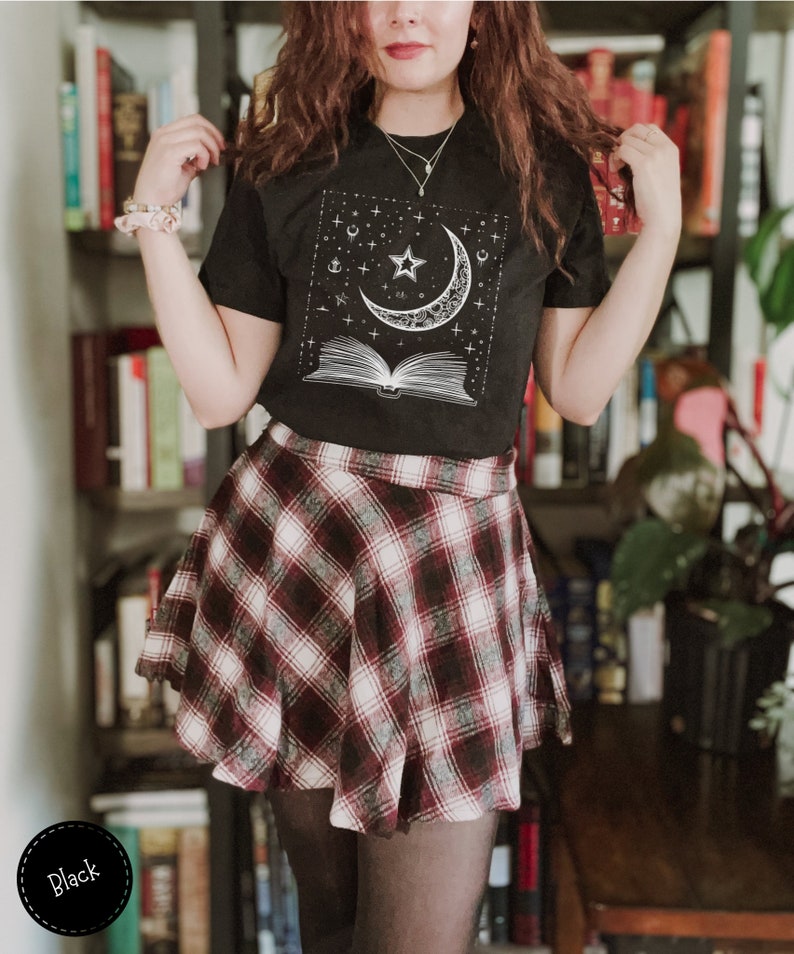 May include: Black t-shirt with a white graphic of a crescent moon, a star, and an open book with stars and other celestial symbols. The shirt is worn with a red and white plaid skirt.