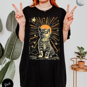 May include: Black oversized t-shirt with a graphic print of a cat looking up at a sun with stars and rays of light. The text "Forest Fables" is in the top right corner of the image.