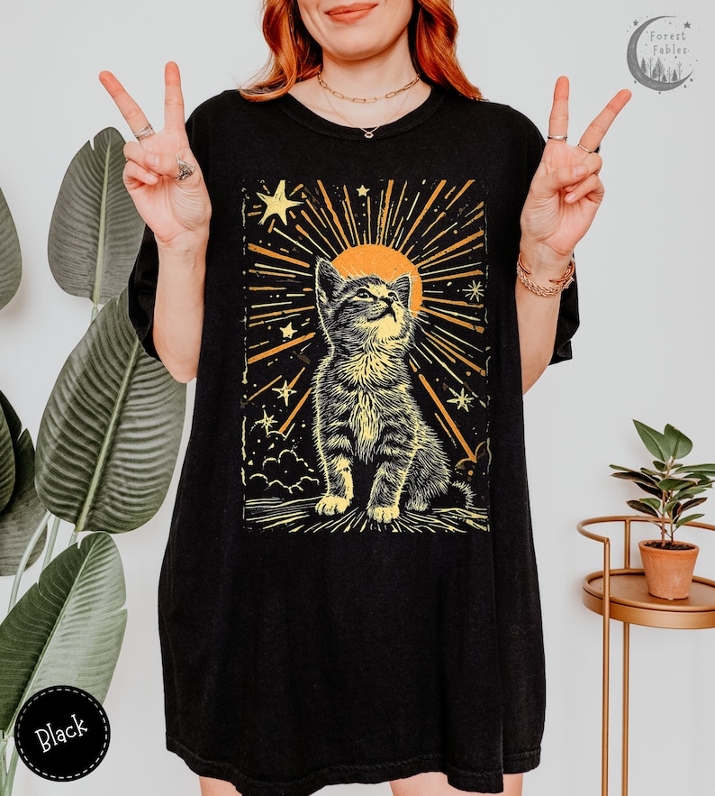 May include: Black oversized t-shirt with a graphic print of a cat looking up at a sun with stars and rays of light. The text "Forest Fables" is in the top right corner of the image.