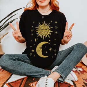 Puede incluir: Una camiseta negra con un dise&ntilde;o de sol y luna dorado. El sol y la luna tienen caras y est&aacute;n rodeados de estrellas. El texto "Forest Fables" est&aacute; en la esquina superior derecha de la imagen.