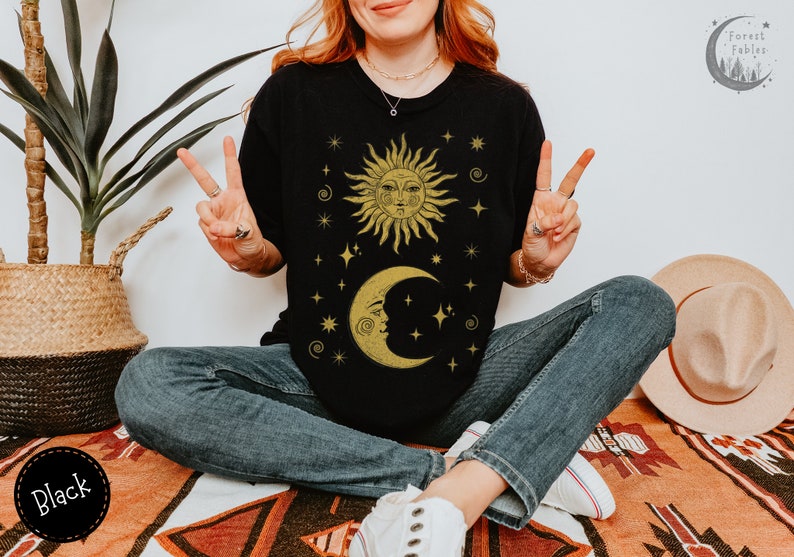 Puede incluir: Una camiseta negra con un dise&ntilde;o de sol y luna dorado. El sol y la luna tienen caras y est&aacute;n rodeados de estrellas. El texto "Forest Fables" est&aacute; en la esquina superior derecha de la imagen.