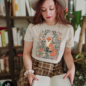 May include: A cream-colored t-shirt featuring a seated fox illustration surrounded by flowers. The fox has orange and white fur. The shirt is paired with a brown plaid skirt. The text "Soft Cream" is in a black circle.