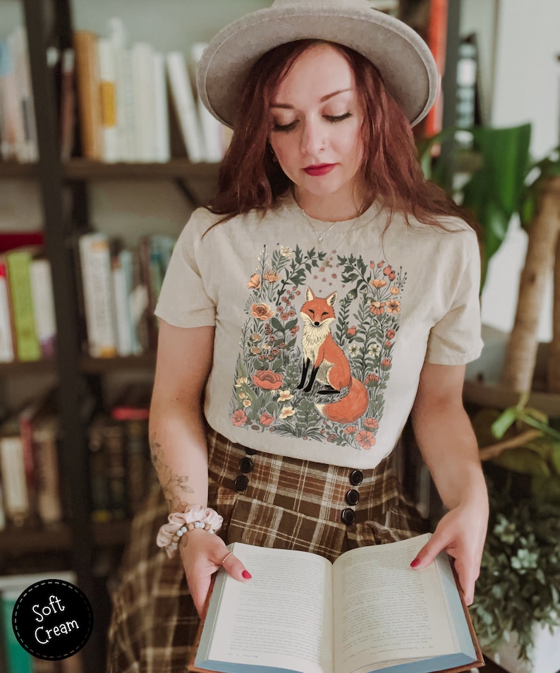 May include: A cream-colored t-shirt featuring a seated fox illustration surrounded by flowers. The fox has orange and white fur. The shirt is paired with a brown plaid skirt. The text "Soft Cream" is in a black circle.