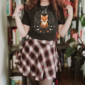 May include: A black t-shirt with a graphic of a fox surrounded by flowers. The person is wearing a red and white plaid skirt.