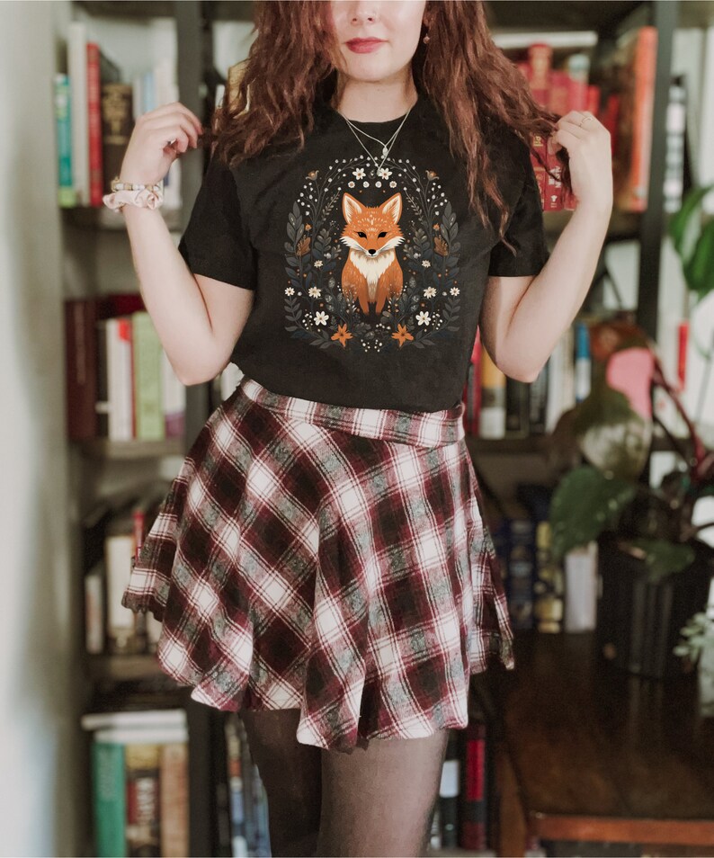 May include: A black t-shirt with a graphic of a fox surrounded by flowers. The person is wearing a red and white plaid skirt.