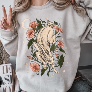 May include: A light grey sweatshirt with a graphic print of a white rabbit jumping through a floral arrangement with a crescent moon and stars. The flowers are pink and orange.