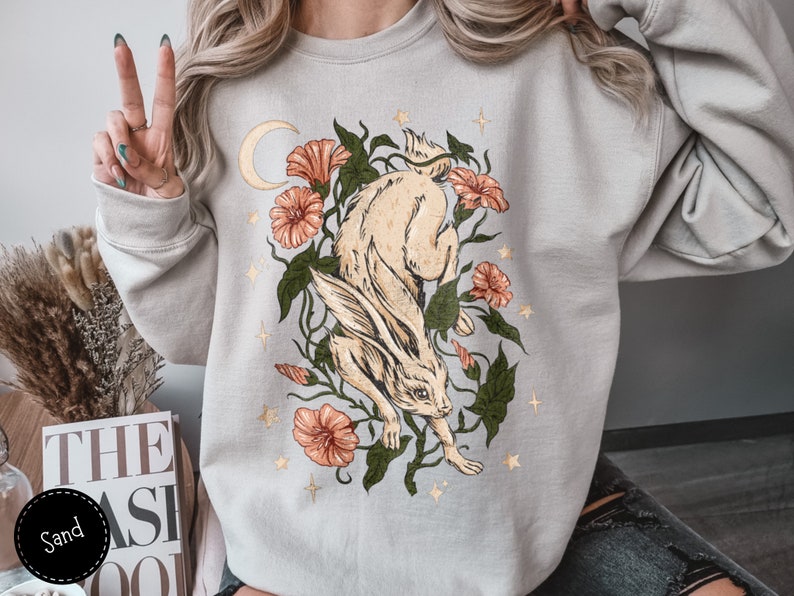 May include: A light grey sweatshirt with a graphic print of a white rabbit jumping through a floral arrangement with a crescent moon and stars. The flowers are pink and orange.