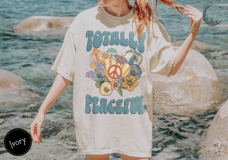 May include: A white t-shirt with a retro graphic design featuring the text "Totally Peaceful" in blue letters with a peace sign and flowers. The shirt is a relaxed fit and has a crew neck.