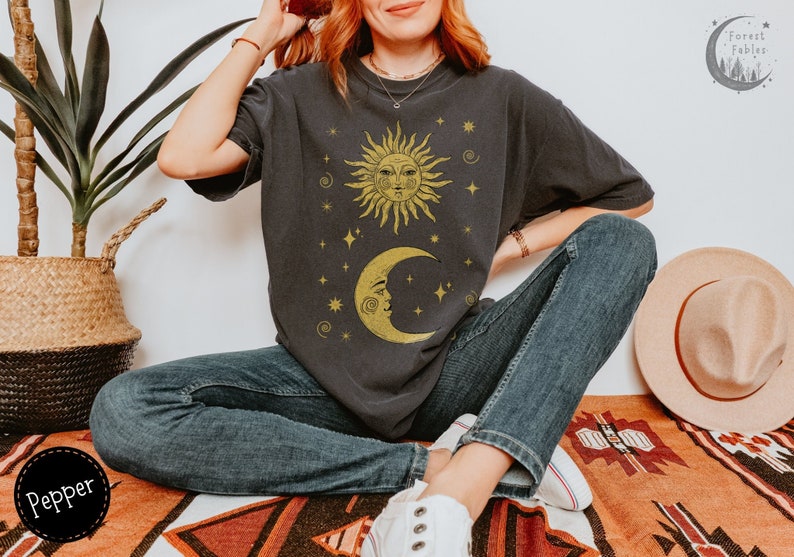 Puede incluir: Una persona lleva una camiseta negra con un dise&ntilde;o de sol y luna dorado. La camiseta tiene un aspecto desgastado y presenta un gr&aacute;fico de estilo vintage. La persona est&aacute; sentada en una alfombra estampada con un sombrero y una planta en el fondo.