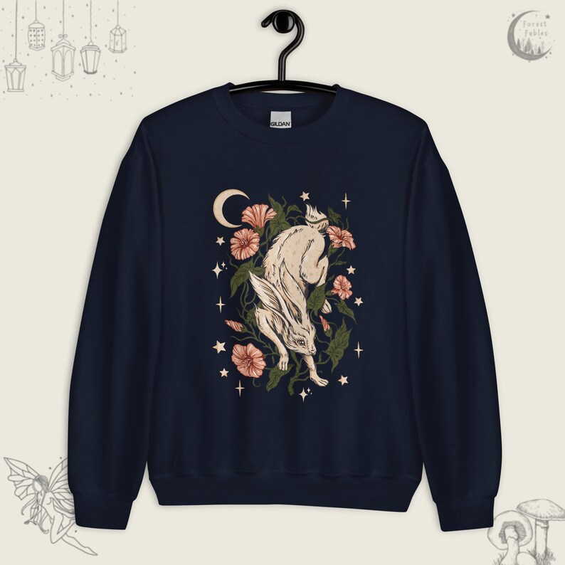 May include: A navy blue sweatshirt with a white rabbit jumping through a floral arrangement. The rabbit is surrounded by pink flowers, green leaves, and a crescent moon. The background is a dark blue with white stars.