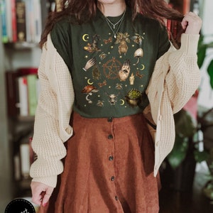 Puede incluir: Una camiseta verde oscuro con un estampado de varios s&iacute;mbolos e ilustraciones de brujer&iacute;a, incluyendo una luna creciente, una mano sosteniendo una rama de romero, un caldero y una calavera de gato. La camiseta la lleva puesta una persona con el pelo largo y casta&ntilde;o y un c&aacute;rdigan blanco.