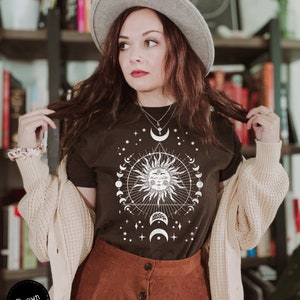 K&ouml;nnte beinhalten: Ein schwarzes T-Shirt mit einem wei&szlig;en Motiv einer Sonne mit einem Gesicht und einem Halbmond. Das Motiv enth&auml;lt auch Sterne und ein Dreieck. Das Shirt wird von einer Person mit braunen Haaren und einem grauen Hut getragen.