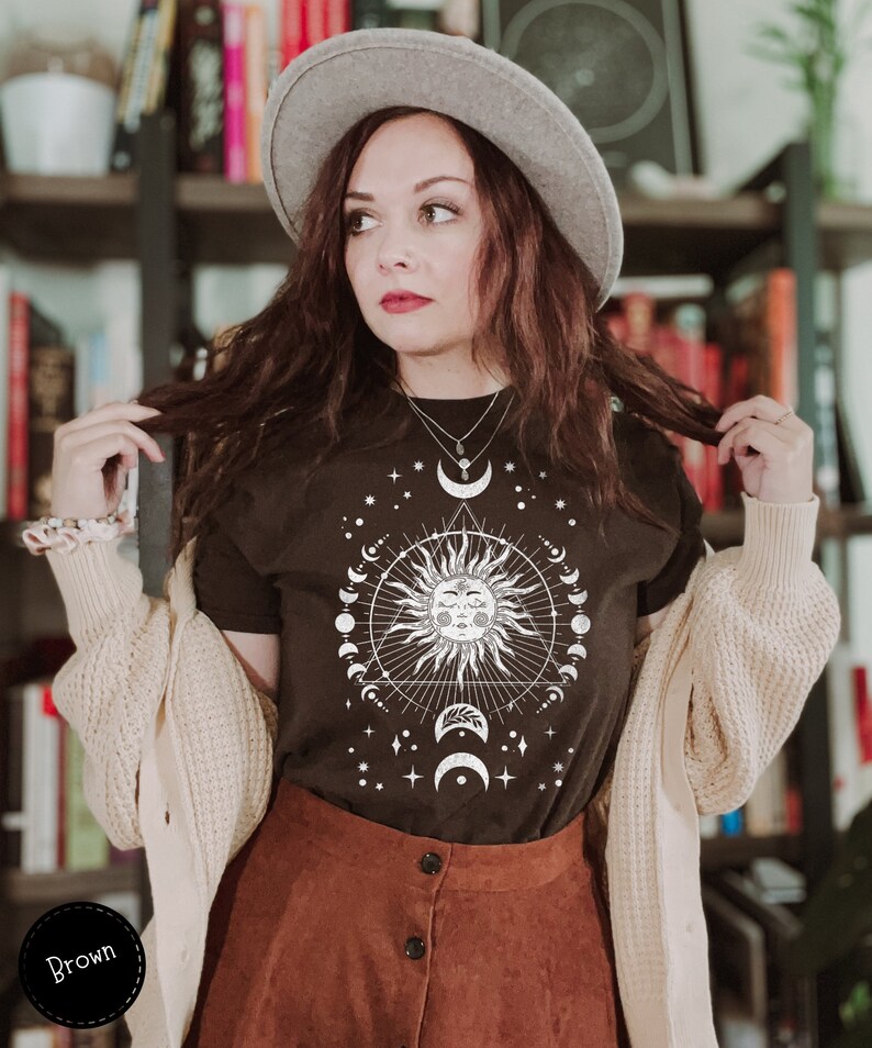 K&ouml;nnte beinhalten: Ein schwarzes T-Shirt mit einem wei&szlig;en Motiv einer Sonne mit einem Gesicht und einem Halbmond. Das Motiv enth&auml;lt auch Sterne und ein Dreieck. Das Shirt wird von einer Person mit braunen Haaren und einem grauen Hut getragen.