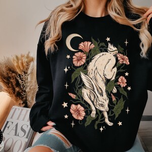 May include: Black sweatshirt with a graphic print of a white rabbit running through a forest with pink flowers and a crescent moon.