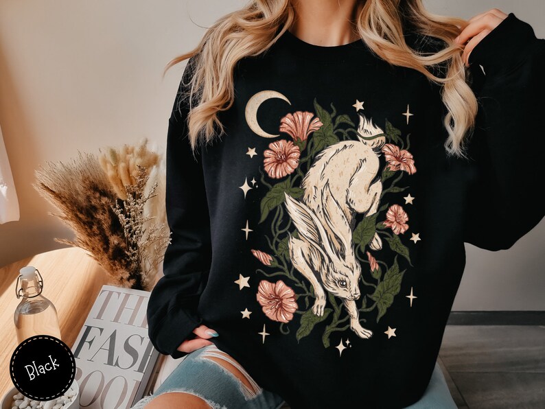 May include: Black sweatshirt with a graphic print of a white rabbit running through a forest with pink flowers and a crescent moon.