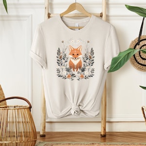 May include: A beige t-shirt with a graphic of a fox surrounded by flowers and leaves. The fox is orange and white with black eyes.
