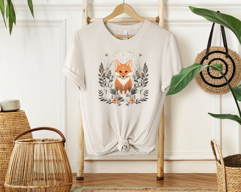 May include: A beige t-shirt with a graphic of a fox surrounded by flowers and leaves. The fox is orange and white with black eyes.