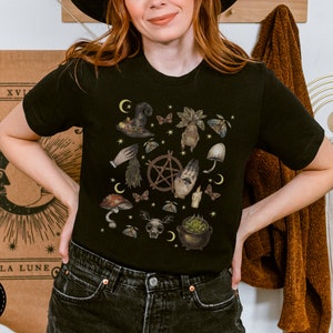 Puede incluir: Camiseta negra con un estampado gr&aacute;fico de un caldero de bruja, setas y otros elementos m&iacute;sticos. El texto "Forest Fables" es visible en la esquina superior derecha de la imagen.