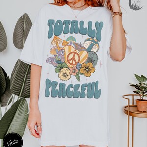 May include: White t-shirt with a retro graphic design featuring the text "Totally Peaceful" in blue with a peace sign and flowers in a heart shape.