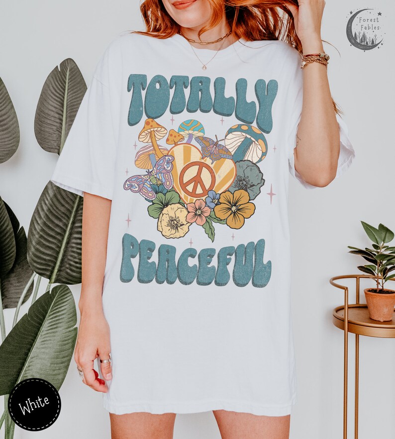 May include: White t-shirt with a retro graphic design featuring the text "Totally Peaceful" in blue with a peace sign and flowers in a heart shape.